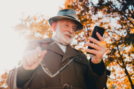 Elderly man outdoors enjoying a bright autumn day while checking his smartphone in a park with...