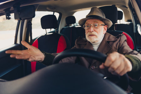 Senior man driving a car on a sunny autumn day wearing a coat and hat, showing expressive gestures...