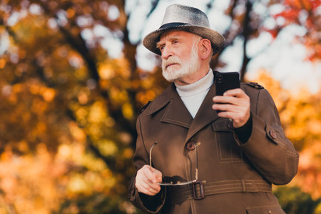 Elderly man in fall attire standing outdoors with phone and glasses in an autumn park surrounded by...