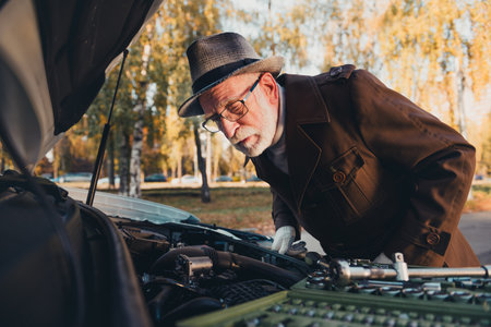 Senior man in an outdoor autumn park inspecting his car engine with tools under the sunlight on a sunny dayの写真素材