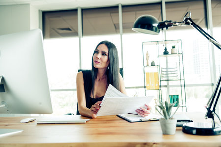 Confident businesswoman analyzing documents at her modern office desk with a computer and elegant...