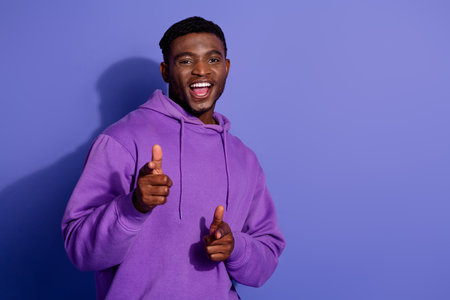 Cheerful young man in purple hoodie gestures playfully on violet background showcasing vibrant...