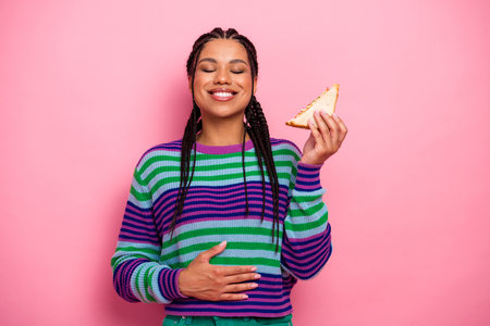 Happy woman with joyful expression enjoys snack holding sandwich, dressed in vibrant striped sweater...