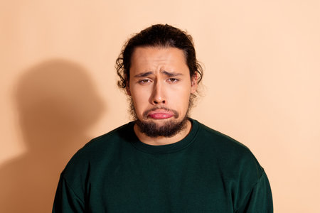 Young man in a green sweatshirt displaying a humorous sad pouty expression against a warm beige backgroundの写真素材
