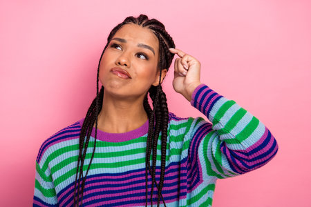 Thoughtful young woman with braided hair wearing a colorful striped sweater posing against a pink...
