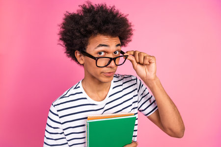 Young male student with glasses holding notebooks in front of pink background