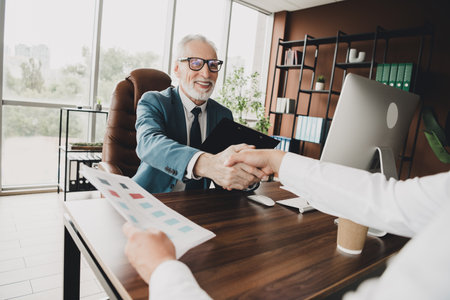Mature businessman in elegant office shaking hands with a colleague, representing professional success and collaboration.の写真素材
