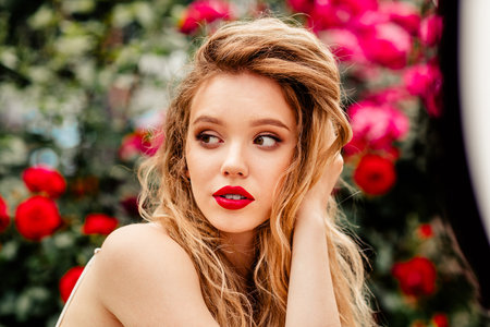 Beautiful woman with bright red lipstick posing outdoors in front of blooming roses on a sunny day