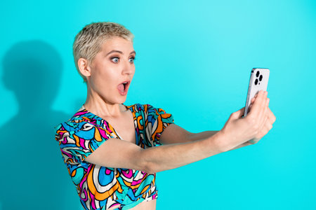 Young woman with a shocked expression taking a selfie on a turquoise background wearing a colorful...