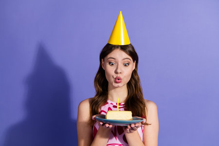 Young woman in party hat blowing candle on slice of cheesecake against purple background, expressing...