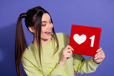 Young woman with long hair holding a heart symbol board against a purple background expressing...