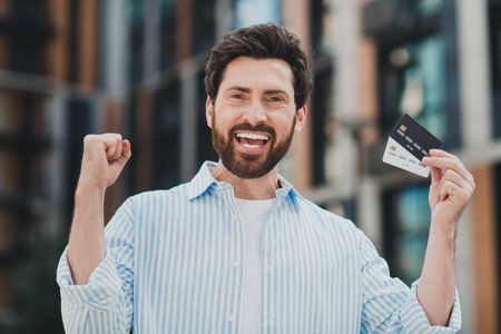 Handsome man in casual attire holding credit cards outdoors with a triumphant smile in an urban city...