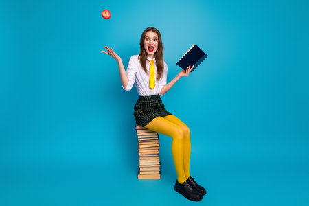 Young student in colorful retro uniform sitting on book, holding a book and an apple against a vibrant blue backgroundの写真素材