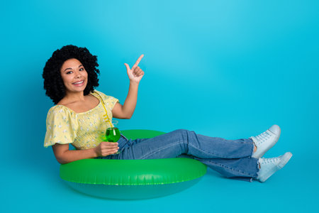 Young woman in casual summer attire sitting on inflatable ring and holding a green drink against...