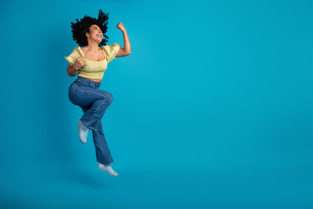 Young woman jumping joyfully on a blue background wearing a yellow top and jeans, expressing...