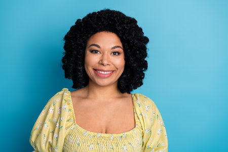 Cheerful young woman with curly hair in a casual yellow outfit posing against a blue background...