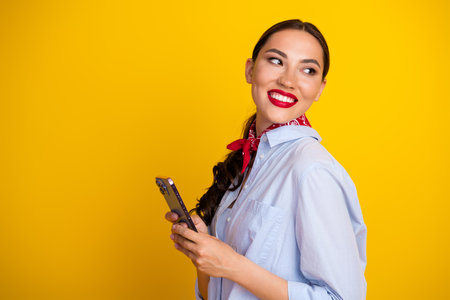 Smiling woman holding a smartphone against a vibrant yellow background, showcasing fashion and...
