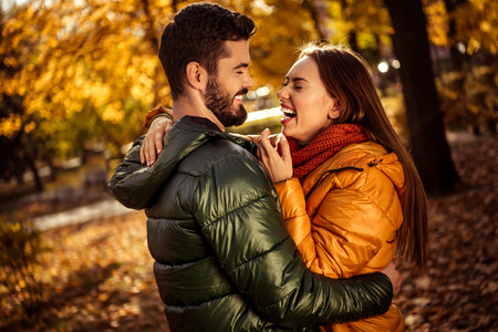 Romantic couple laughing together in an autumn park surrounded by vibrant foliage and warm sunlight embracing seasonal joyの写真素材