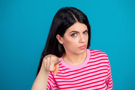 Portrait of a serious young woman pointing forward against a vibrant blue background while wearing a stylish striped shirtの写真素材