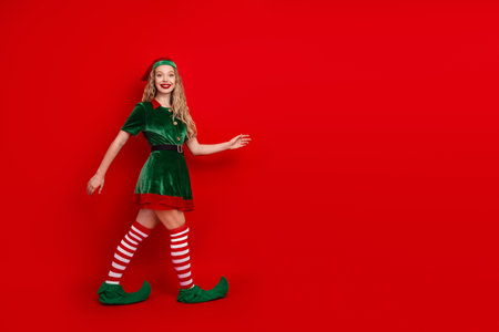 Young woman dressed as a cheerful Christmas elf posing on a vibrant red background, embodying...