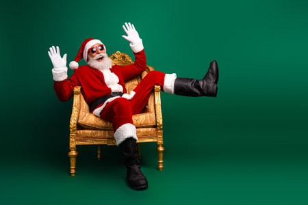 Santa Claus in festive costume sitting joyfully on golden chair with playful pose against vibrant green backgroundの写真素材