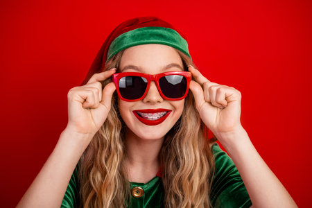 Trendy young woman in festive elf costume with red sunglasses against vibrant background celebrating holiday joy.の写真素材