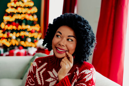 Smiling woman in cozy Christmas sweater enjoying festive holiday atmosphere with illuminated tree in cozy room interiorの写真素材