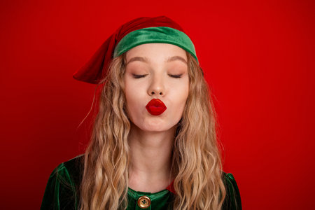 Festive young woman dressed as an elf happily kissing in front of a vibrant red background...