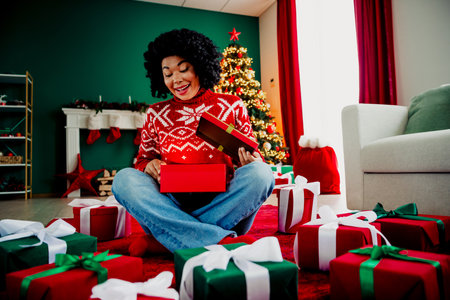 Smiling woman opening gift box surrounded by presents under decorated Christmas tree in cozy living...