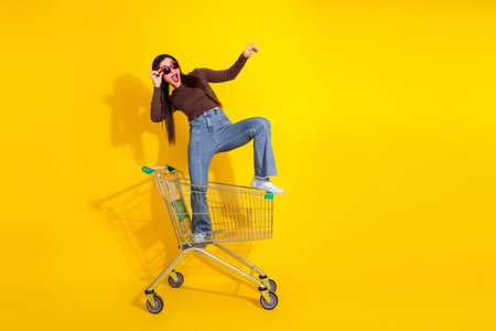 A cheerful young woman posing stylishly on a shopping cart against a vibrant yellow backdrop,...