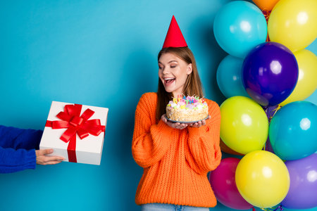 Adorable young woman in an orange sweater holding a birthday cake while receiving a gift amid...