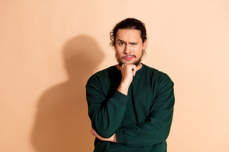 Confident young man in green sweatshirt posing against beige background with thoughtful expression...