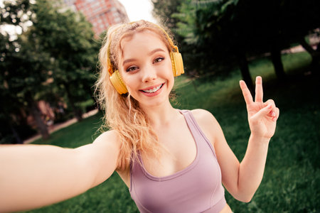Young woman practicing fitness outdoors, wearing yellow headphones, showing peace gesture, in bright...