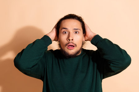 Portrait of a stylish young man in a green sweatshirt expressing surprise in front of a beige...