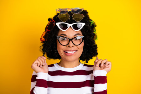 Playful young woman with curly hair and glasses posing against a vibrant yellow background...