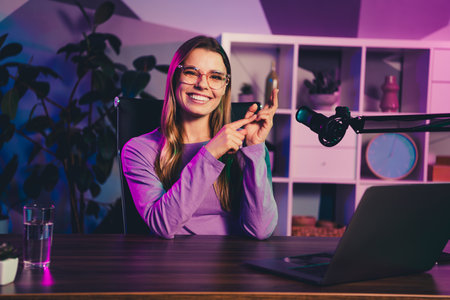 Radio host hosting a live podcast at a stylish home office desk with microphone and laptop against vibrant neon lightingの写真素材