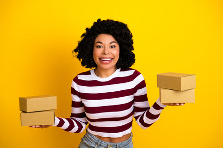 Smiling young woman holding cardboard boxes in both hands against a yellow background wearing a...