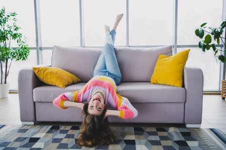 Young woman in a colorful sweater relaxing upside down on a sofa with a cheerful expression in a cozy living room settingの写真素材