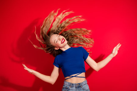 Joyful young woman with blonde hair dancing energetically in a blue top against a vibrant red...