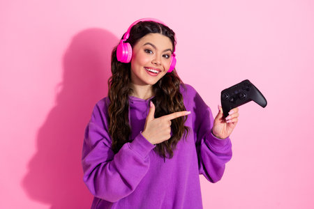 Smiling young woman in purple sweatshirt holding black gaming controller against pink background,...