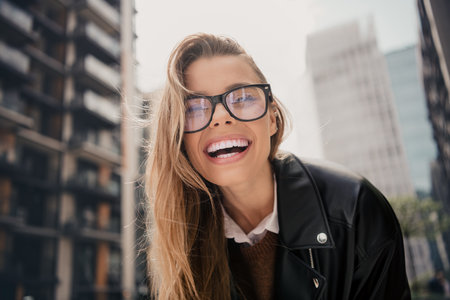 Cheerful young woman with glasses and blonde hair enjoying an urban cityscape on a sunny day in a...