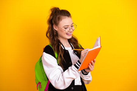 Cheerful young female student taking notes with a bright smile against a vibrant yellow background...