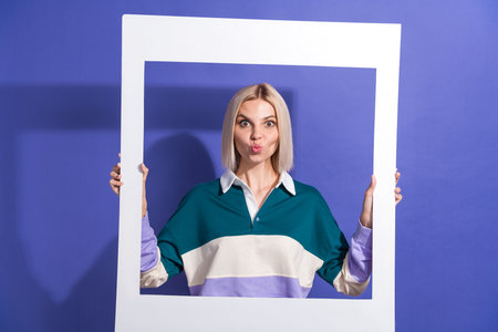 Portrait of a young blonde woman posing playfully with a frame on a purple background