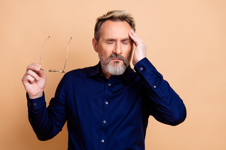 Mature businessman holding glasses with hand on head, expressing concentration and stress against beige backgroundの写真素材