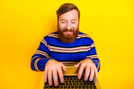Cheerful bearded man in a colorful striped sweater using a laptop against a vibrant yellow...