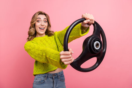 Cheerful woman holding a steering wheel against pink background wearing stylish knit sweater and denim jeansの写真素材