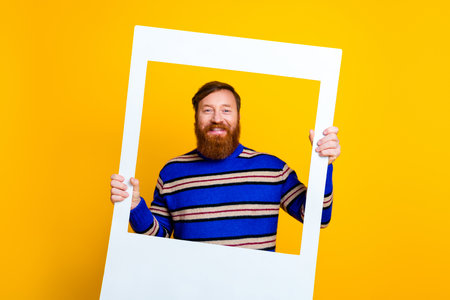 Cheerful man with red hair smiling while holding a photo frame on a bright yellow background