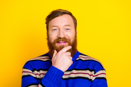Handsome bearded man in a stylish striped sweater on a vibrant yellow background with thoughtful gesture and smileの写真素材