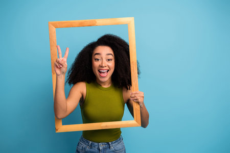 Joyful young woman with natural curls posing in wooden frame against solid blue background, showing...