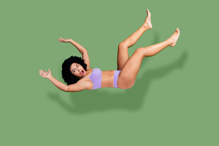 Confident woman in purple lingerie posing with a surprised expression in mid-air against a green backgroundの写真素材